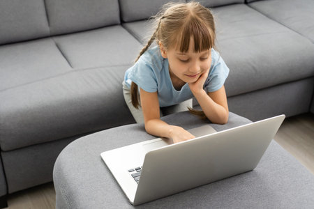 Bored little girl with laptop at home. Cute child doing homework on computer. Modern online...