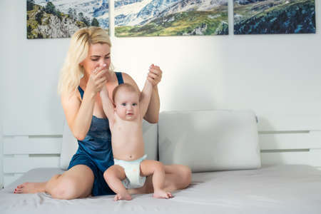 Mother and child on a white bed. Mom and baby boy in diaper playing in sunny bedroom. Parent and little kid relaxing at home. Family having fun together. Bedding and textile for infant nurseryの写真素材