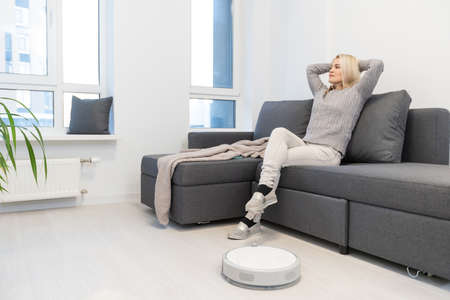 Robotic vacuum cleaner cleaning the room while woman relaxing on sofa. Woman controlling vacuum with remote control.の写真素材