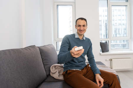 Weekend leisure activities. Millennial Caucasian man with remote control switching channels on TV at home. Happy young guy watching interesting program on television indoorsの写真素材