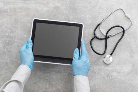 Doctor hand holding tablet with blank screen and stethoscope on grey table, copy space, top viewの写真素材