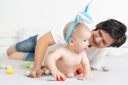 Mother and child with colorful eggs. Mom and baby with bunny ears. Parent and kid play indoors in...