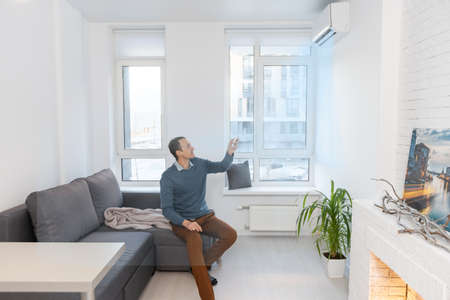 Young Man On Sofa Operating Air Conditioner With Remote Control.の写真素材