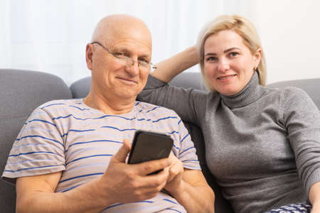 Smiling older father with adult daughter using phone together, having fun, watching video in social network or shopping online, caring young woman teaching mature dad or grandfather to use smartphoneの写真素材