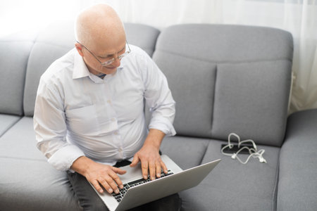 Elderly bald man in white shirt and black pants correcting glasses and working on laptop while...