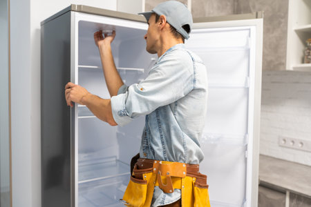 young repairman with tool belt looking at broken refrigeratorの写真素材