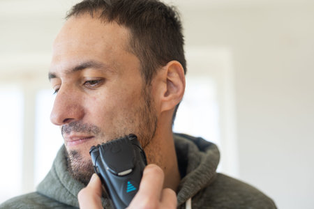 Handsome young man shaving with electric razor over gray background.