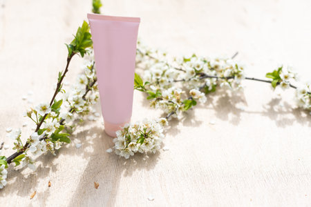 Blank white cosmetics tube and spring flowering tree branch with white flowers on pastel background....