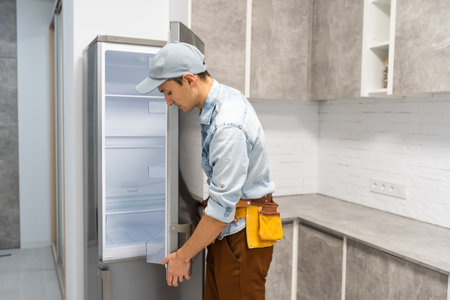 young repairman with tool belt looking at broken refrigeratorの写真素材