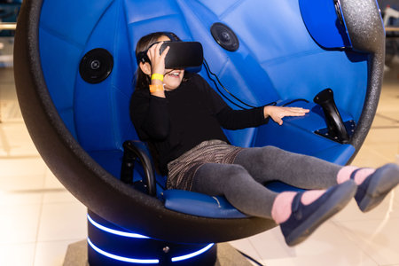 A little girl sitting in a gaming cockpit and holding the race steering wheel to play a racing...