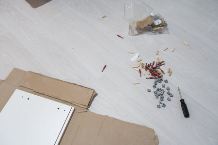 Parts of unfinished furniture, metal screws and tools lying on the floor with instruction manual for...