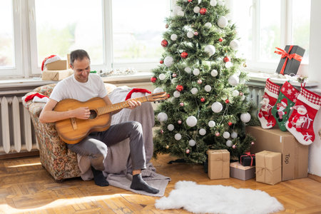 Happy young man is playing guitar. Guy is looking happily and carefree. Male in festive hat alone celebrating Christmas or new year. Christmas tree with garland in backgroundの写真素材