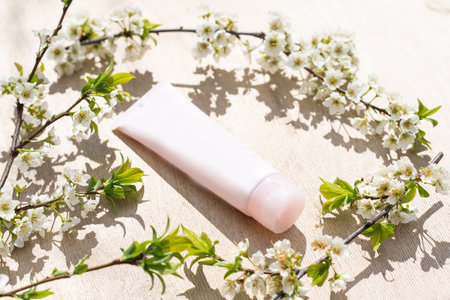Blank white cosmetics tube and spring flowering tree branch with white flowers on pastel background....