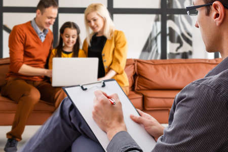 Hand of a professional family psychotherapist writing notes in front of a family with a child during a consultationの写真素材