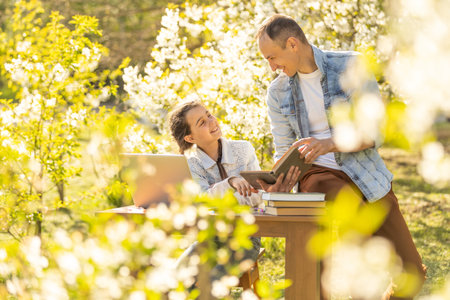 A girl and a young father are sitting at a laptop and studying in a flowered garden. Against the...