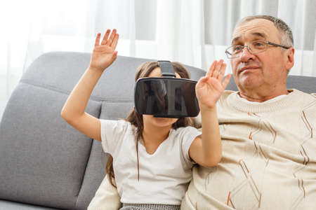 senior Caucasian man at home in the living room on the couch, grandfather and granddaughter with virtual glasses.の写真素材