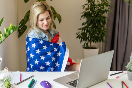 Happy young female student hold flag of USA, studying with laptop with blank screen in living room interior. International education at home, lesson remote, website due covid-19 quarantineの写真素材
