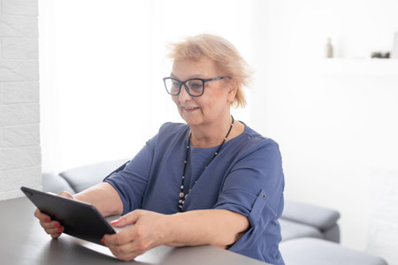 Merry mature middle-aged woman spends leisure time at home using a digital tablet. Cheerful modern...