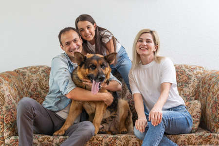 Beautiful young parents, their cute little daughter looking at camera and smiling, sitting with their cute dog on sofa at homeの写真素材