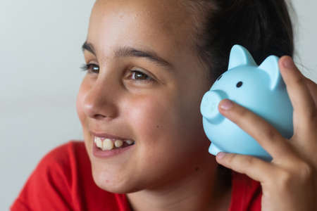Smiling little girl posing with a piggy bank in her hands, standing against a blue background in a studio with free space. Family savingsの写真素材