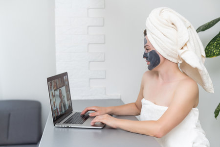 Young woman with towel on head and black facial clay mask sitting at home and using laptop computer . Staying at home, quarantine, distance learning, online education, technology, modern lifestyleの写真素材