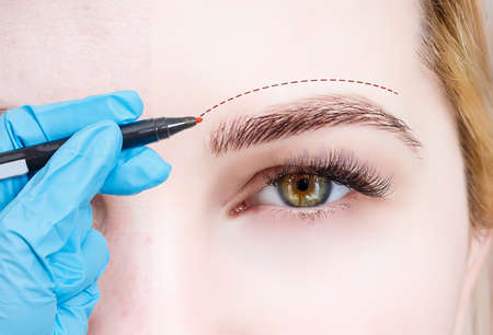 Plastic surgeon drawing dashed line under eye of girl. Hand in blue glove holding pencil. Plastic surgery, beauty portrait, closeup.の写真素材