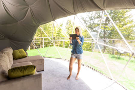 Young woman controlling home light with a digital tablet in the glamping dome tent. Concept of a smart home and light control with mobile devicesの写真素材