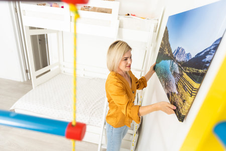 Modern Home Interior And Domestic Decor. Smiling young woman hanging painting, putting photo picture...