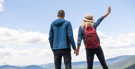 Rear view of hiking couple with backpack standing together on hill top enjoying beautiful landscape. Man and woman outdoors on hiking standing on a rockの写真素材