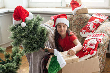 little girl makes an artificial Christmas tree wear santa hat indoorsの写真素材