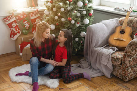 Beautiful mother and daughter sitting by nicely decorated Christmas tree, having fun at home on Christmas dayの写真素材