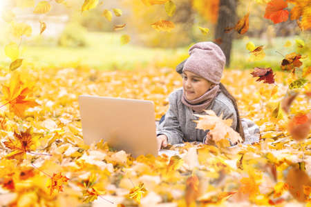 Cute little girl holds an orange maple leaf near the face. Pretty blond girl wearing beige coat, knitted cap and scarf has fun in autumn park, close-up.の写真素材