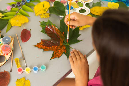 little girl artist with a brush and paints in her hands in autumn idraws a landscape with leaves on canvasの写真素材