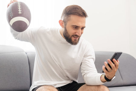 American football fan holding rugby ball and watching match on smartphone