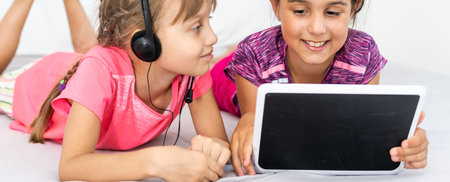 Two cute little girls are using a digital tablet and smiling while lying on bed in childrens room