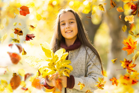 Cute little girl holds an orange maple leaf near the face. Pretty blond girl wearing beige coat, knitted cap and scarf has fun in autumn park, close-up.の写真素材
