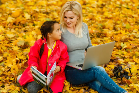 Happy smiling mother and child sits together looking on book or tablet pc in autumn parkの写真素材