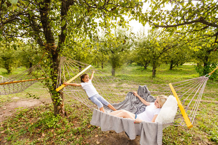 Beautiful young mother and daughter laying down and relaxing together on a hammock during a sunny summer day on holiday home garden. Family relaxing outdoors, healthy and wellness lifestyle.の写真素材