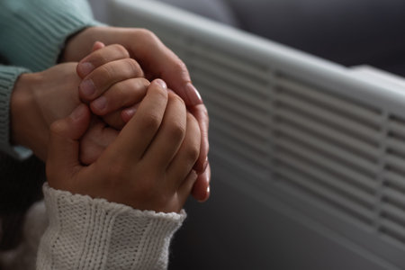 Mother and child warming hands near electric heater at home, closeup.の写真素材