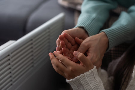 Mother and child warming hands near electric heater at home, closeup.