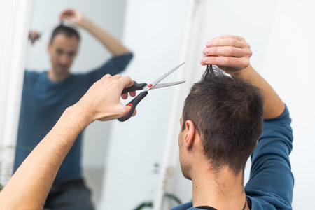 Caucasian shaggy man trying to make a haircut by himself with scissors being on quarantine during pandemic of covid-19の写真素材