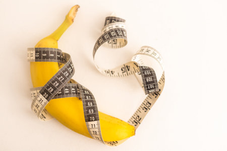 Regime, healthy food and weight management concept. Centimeter ruler spinned around fresh fruit. Yellow tape around banana isolated on light background. Banana with tape for measuring figure.の写真素材