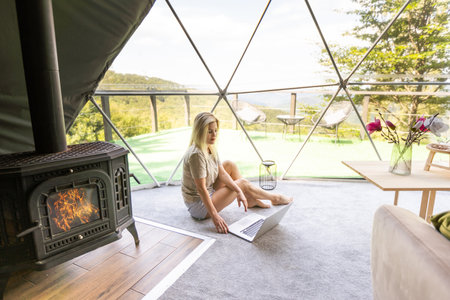 Woman working on laptop geo dome tents. Green, blue background. Cozy, camping, glamping, holiday, vacation lifestyle concept. Outdoors cabin, scenic background.の写真素材