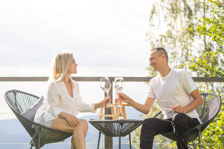 Happy couple sit on wicker chairs at outdoor terrace in the mountains and drink wine. Romantic time together.の写真素材