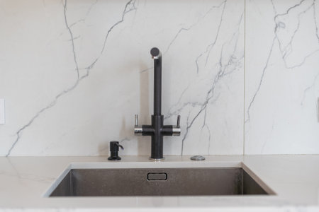A kitchen sink detail with a black faucet, white cabinets, and marble countertopの写真素材