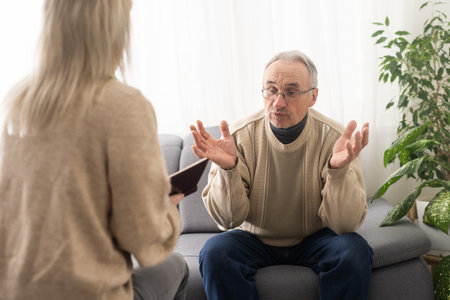 Senior man patient and young woman caregiver medical worker in uniform hold clipboard noting personal information talking listens client telling about health complaints, care support nursing conceptの写真素材