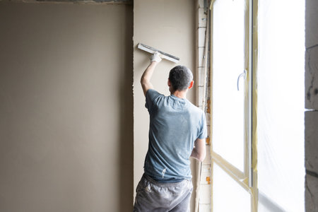 Construction worker wearing worker overall with wall plastering tools renovating apartment house. Plasterer renovating indoor walls and ceilings with float and plaster. Construction finishing works.の写真素材