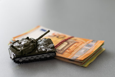 plastic model toy tanks on a pile of euro banknotes against wooden backgroundの写真素材