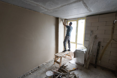 Construction worker wearing worker overall with wall plastering tools renovating apartment house. Plasterer renovating indoor walls and ceilings with float and plaster. Construction finishing works.の写真素材