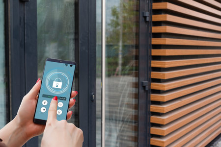 Female hand holding black mobile smart phone with smart home application on the screen. Blurred...